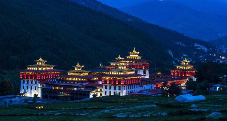 Night view of Tashichho Dzong illuminated in red and gold lights against dark mountains.