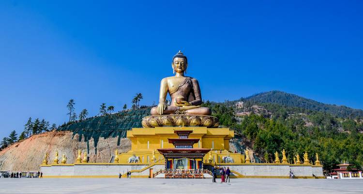 Gigantic golden Buddha Dordenma statue towering above visitors on a wide plaza with forested hills behind.