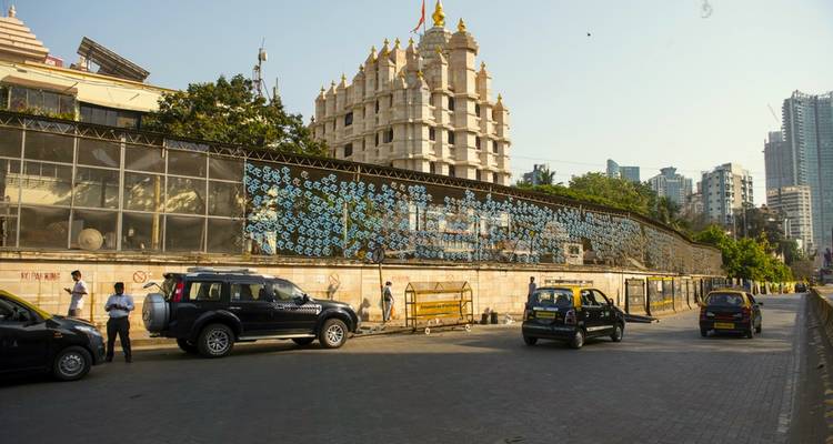 Escena callejera con vehículos y visitantes afuera de un templo Hindu blanco y rascacielos urbanos detrás.