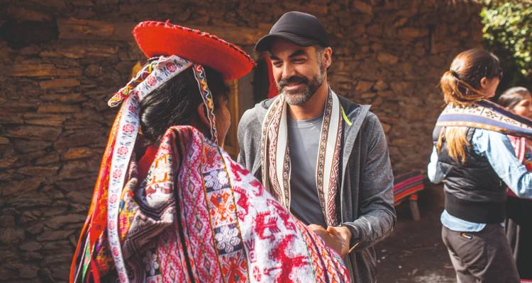 A traveller warmly greets a local woman in vibrant traditional Andean attire outside a stone house.
