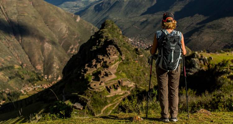 A hiker with trekking poles surveys dramatic Incan ruins and the Sacred Valley from a mountain viewpoint.