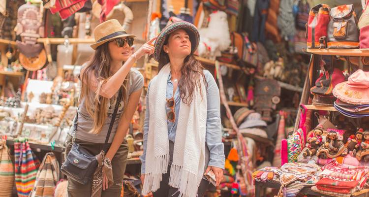 Two women browse colorful handmade hats and textiles in a bustling Andean craft market.