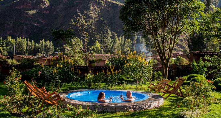 A couple relax with drinks in a circular outdoor hot tub surrounded by lush gardens and mountains.