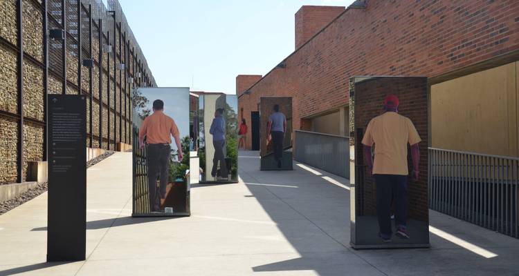 Paseo del museo al aire libre con paneles de fotos a tamaño real de personas contra arquitectura moderna de ladrillo y gaviones en un día soleado.
