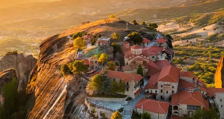 Aerial view of Meteora’s cliff-top monasteries bathed in golden sunset light over rolling Thessalian plains.