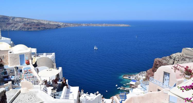 Santorini cliff-side village with white houses overlooking expansive blue sea dotted with a small sailboat