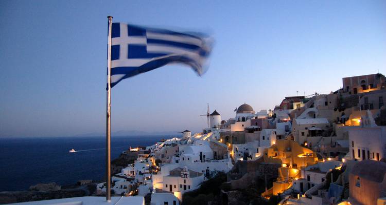 Greek flag waves over the illuminated white houses of Oia perched on cliffs at dusk