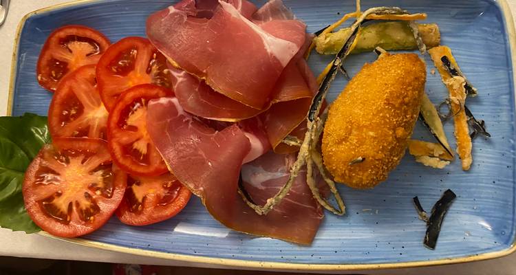 Assiette antipasto italienne présentant des tranches de tomate fraîche, du prosciutto, une croquette frite et des légumes tempura sur une céramique bleue.