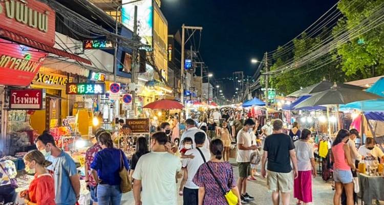 Mercado nocturno tailandés lleno de gente, con letreros brillantes, sombrillas y puestos de comida bajo luces de cadena.