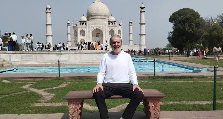 Een glimlachende man zit op een bankje voor de reflectievijver van de Taj Mahal, met bezoekers die achter hem rondlopen.