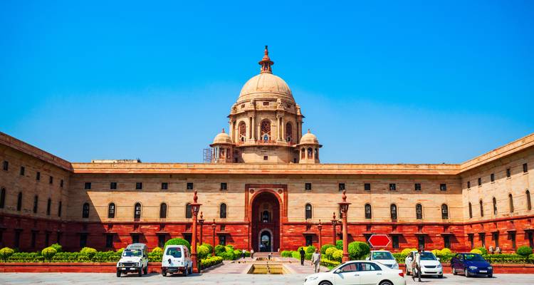 Het indrukwekkende Secretariaat-gebouw van Nieuw-Delhi met zijn centrale koepel kijkt uit op een voorplein vol geparkeerde auto's onder een strakblauwe hemel.