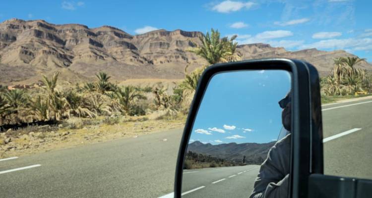 A desert highway and date palms reflect in a 4×4 side mirror with rugged Moroccan mountains ahead.