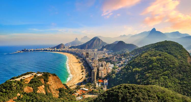 Umfassender Blick auf den Strand von Copacabana, der sich zwischen bewaldeten Hügeln und türkisfarbenem Meer bei Sonnenuntergang erstreckt.
