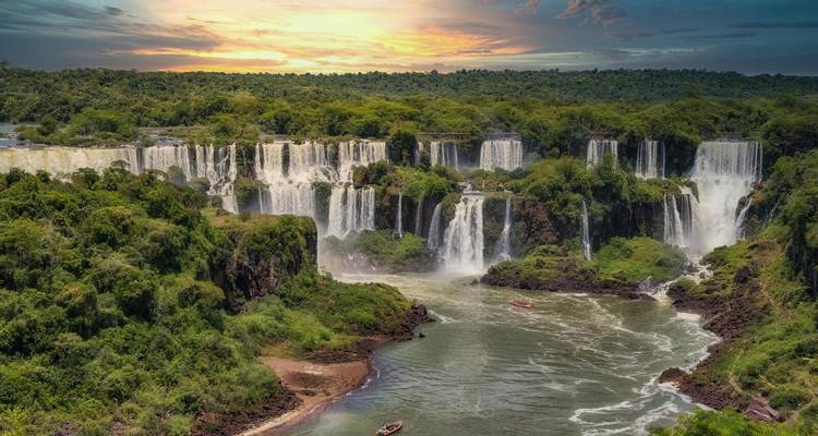 Breiter Panoramablick auf die Iguazu-Fälle bei Sonnenuntergang mit Booten auf dem Fluss darunter.