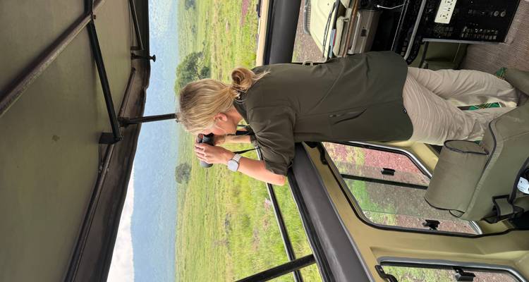 Frau, die in einem Safari-Jeep steht und mit Ferngläsern die Grassteppe absucht.