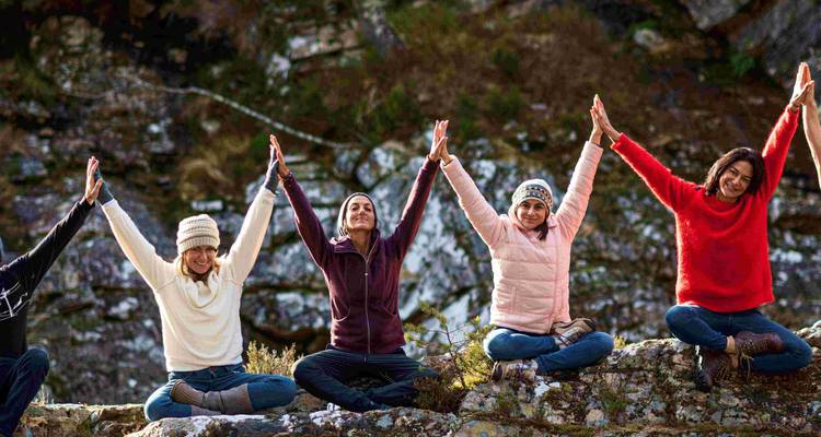 Grupo de personas sentadas en rocas al aire libre levantando los brazos al mismo tiempo durante una sesión de yoga.
