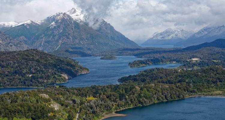 Lac bleu serpentant à travers des montagnes forestières avec de la neige sur les hauts sommets et des nuages au-dessus.