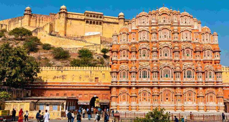 Die verzierte rosa Fassade des Hawa Mahal von Jaipur steht neben den gelben Mauern des Amber Fort unter einem strahlend blauen Himmel, während Touristen darunter spazieren.