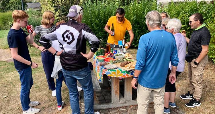 Un rassemblement pique-nique décontracté avec de la nourriture et des boissons disposées sur une nappe colorée tandis que des amis se tiennent debout en discutant à l'extérieur.