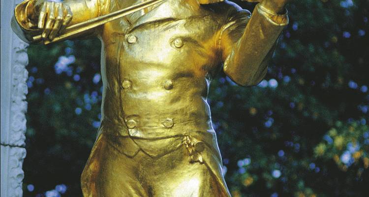 Primer plano de la estatua dorada de Johann Strauss tocando violín contra un fondo frondoso.