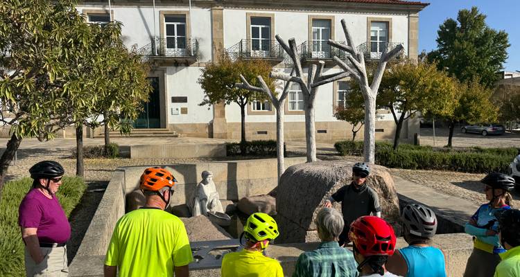 Des cyclistes portant des casques écoutent un guide à côté de sculptures d'arbres modernes dans une place de ville portugaise.