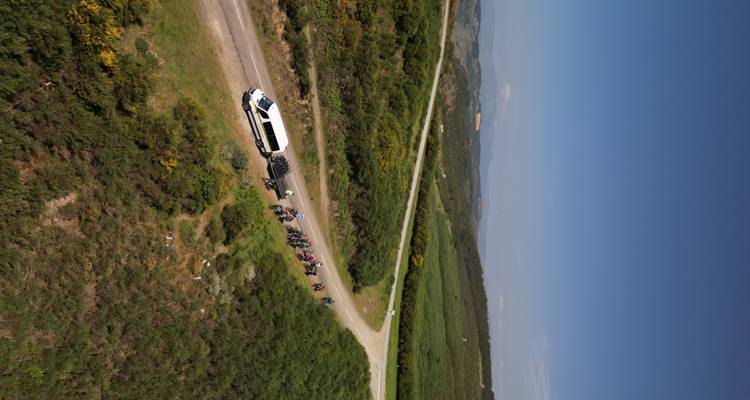 Vue aérienne d'un minibus blanc et d'un groupe de cyclistes circulant sur une route de terre qui serpente à travers des collines verdoyantes ondulantes sous un ciel bleu dégagé.