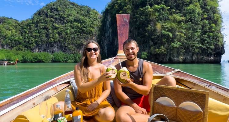 Un couple trinque avec des noix de coco sur un bateau à queue longue naviguant dans des eaux émeraude en passant devant des falaises calcaires.