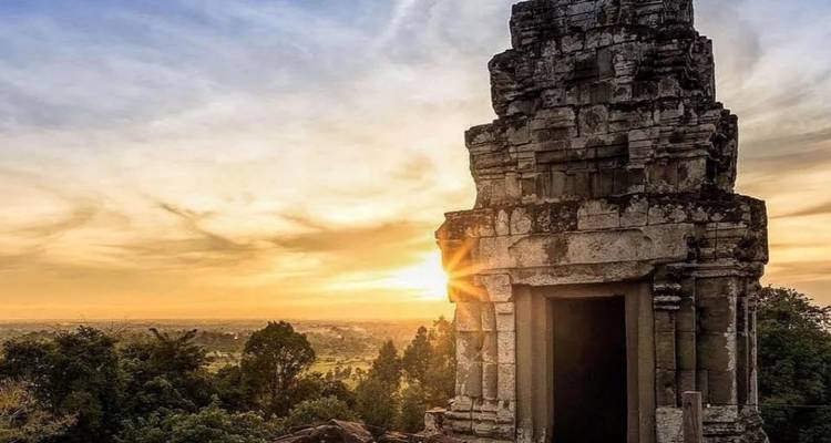 Tour de temple en pierre se détachant en silhouette contre un coucher de soleil doré et chaud sur les plaines cambodgiennes.