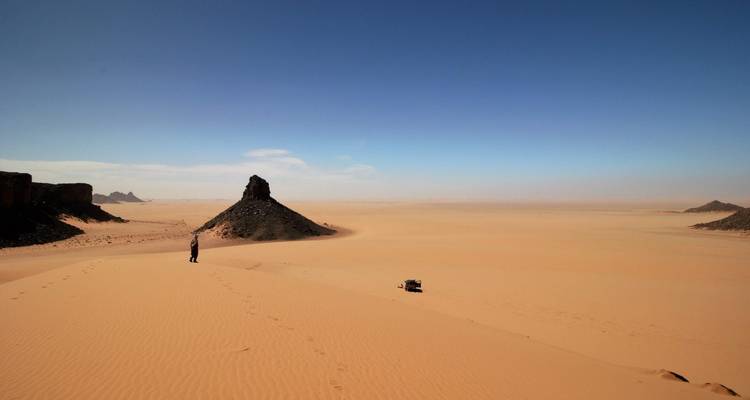 Lone traveller stands in an endless sandy expanse with a dark volcanic hill in the distance.