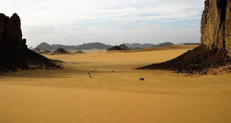 Desert valley bordered by rocky walls with distant off-road vehicles crossing the dunes.