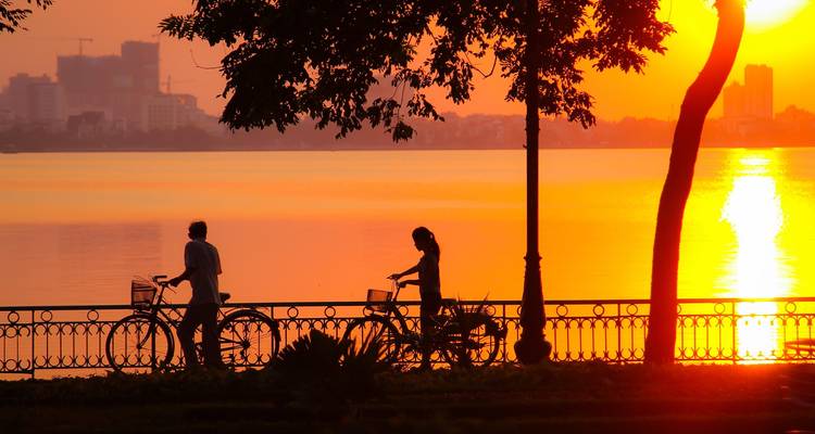 Silhouettes de deux cyclistes poussant des vélos le long d'une promenade au bord d'un lac au coucher de soleil orange éclatant.