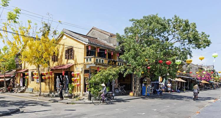 Coin ensoleillé dans la vieille ville de Hoi An avec des maisons patrimoniales jaunes, des lanternes suspendues et des cyclistes.