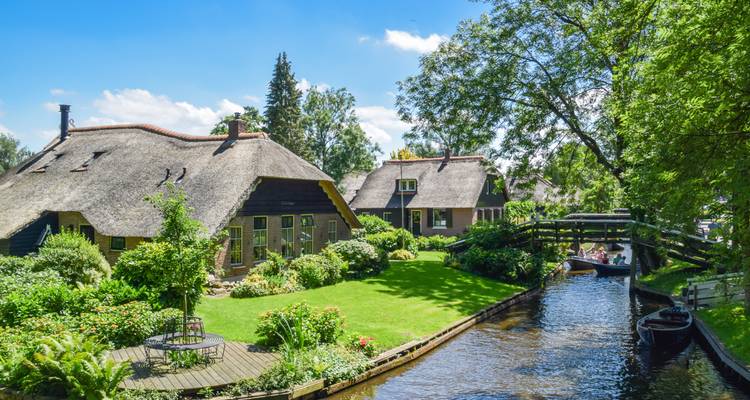 Grüne Kanalszene in Giethoorn mit gepflegtem Rasen, gewölbter Holzbrücke und strohgedeckten Häusern unter blauem Himmel.