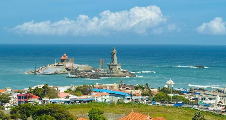 Wide coastal view of the Vivekananda Rock Memorial and Thiruvalluvar Statue offshore with blue sea and town below.
