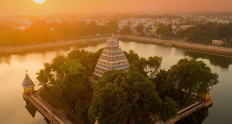 Aerial sunset view of a temple island surrounded by a sacred tank and cityscape beyond.