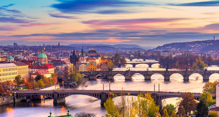 Coucher de soleil panoramique sur Prague avec plusieurs ponts traversant la rivière Vltava et des reflets de ciel colorés.
