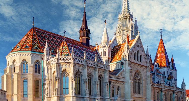 Extérieur de l'église Matthias à Budapest avec son toit aux carreaux colorés sous un ciel partiellement nuageux.