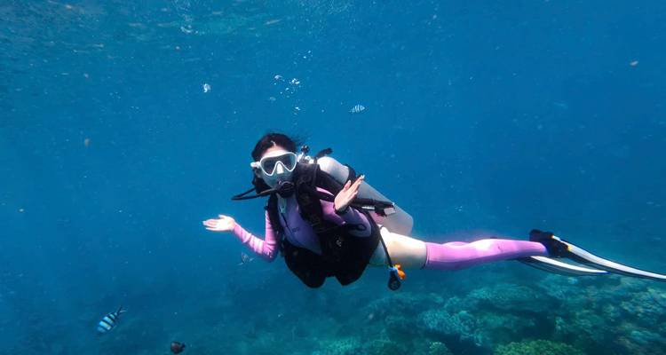 Buceador posando de forma juguetona bajo el agua con un arrecife de coral debajo y peces pequeños alrededor.