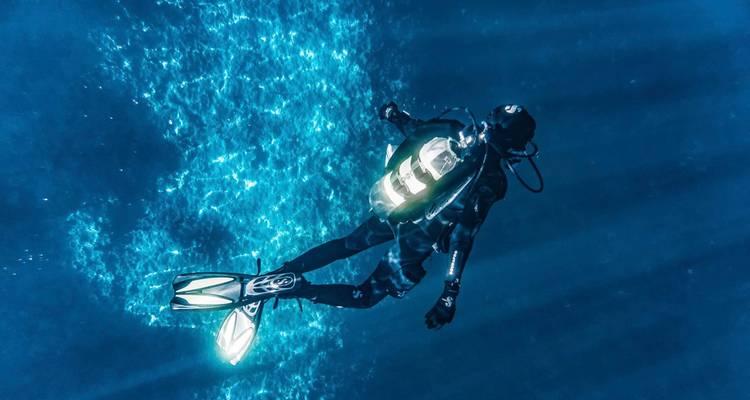 Buzo de buceo con luz de fondo descendiendo a través de rayos de luz en agua azul profunda.