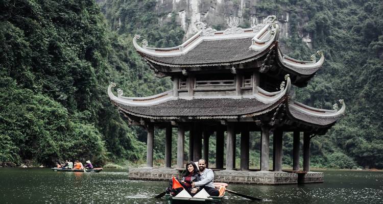 Pavillon traditionnel au bord du lac entouré de montagnes verdoyantes avec un couple souriant dans une sampan à Trang An.