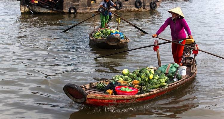 Un vendeur coiffé d'un chapeau conique fait naviguer un bateau en bois chargé de produits à travers les eaux calmes d'un fleuve.
