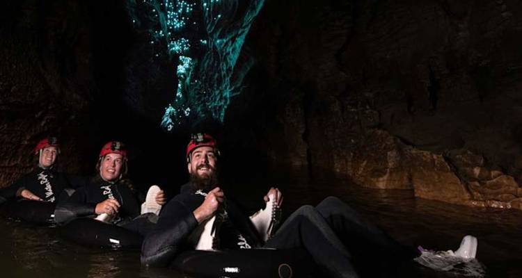 Des aventuriers vêtus de combinaisons étanches flottent sur des chambres à air à travers une grotte sombre illuminée par la lueur bleu-vert des vers luisants au-dessus.