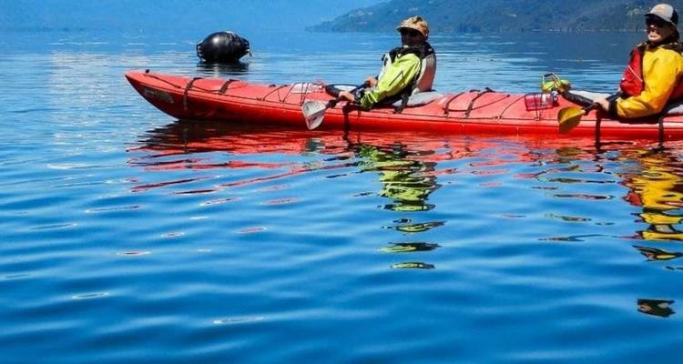 Deux kayakistes se détendent sur un kayak tandem rouge sur une eau de lac bleue et miroir avec des montagnes au-delà.