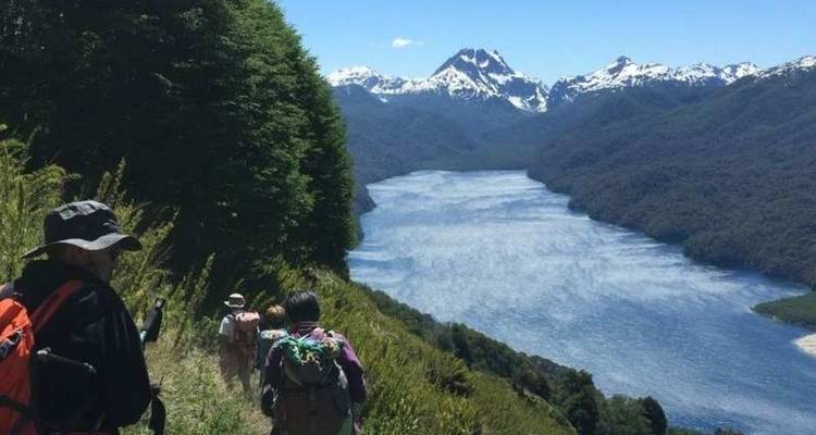 Les randonneurs suivent un sentier alpin étroit surplombant à grande hauteur un long lac aux allures de fjord, avec des pics enneigés au-delà.