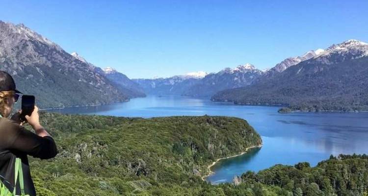 Un voyageur photographie un vaste lac bleu et des montagnes boisées sous un ciel éclatant.