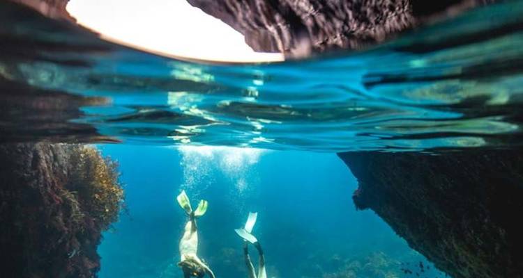 Deux plongeurs en apnée glissent dans une eau bleu clair entre des parois rocheuses imposantes, la lumière du soleil filtrant d'en haut.