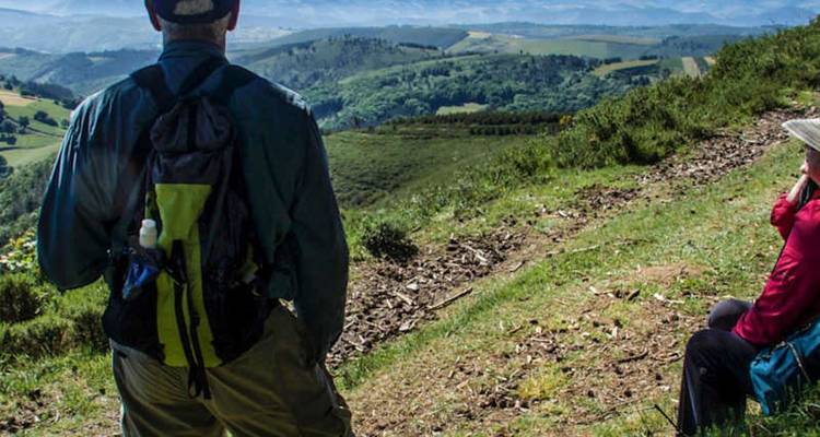 Dos peregrinos descansan en una cresta de sendero cubierta de pasto admirando los amplios valles verdes a lo largo del Camino Primitivo.