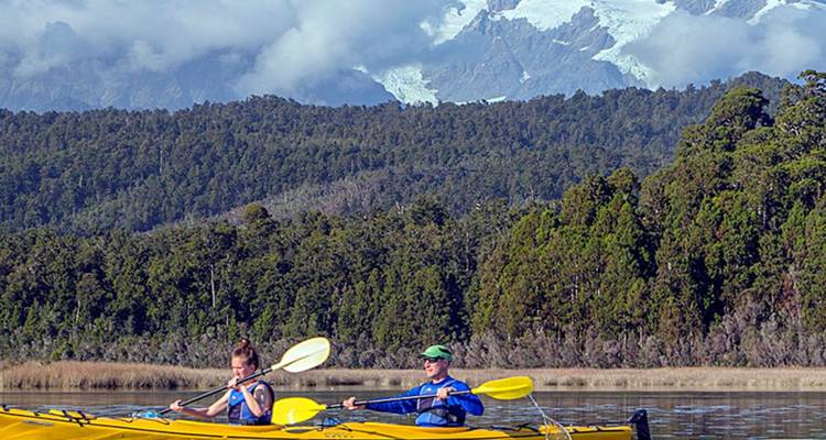 Deux kayakistes pagaient sur un lac calme entouré d'une forêt dense avec des montagnes enneigées s'élevant majestueusement en arrière-plan.