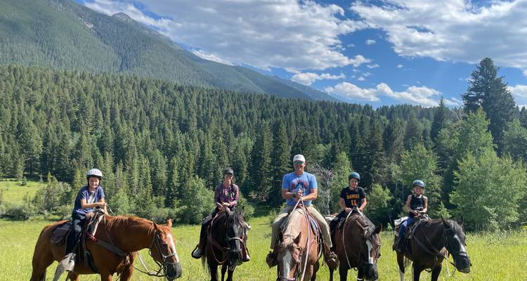 Un groupe familial chevauche à travers un pré verdoyant, avec en arrière-plan une dense forêt de pins et des montagnes.