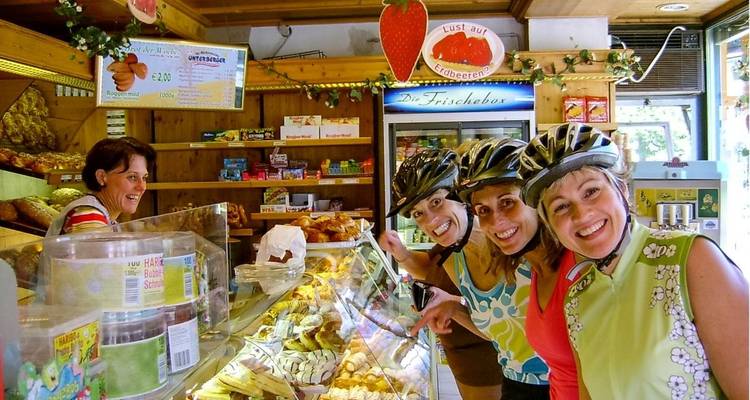 Ciclistas con casco sonríen dentro de una panadería austriaca tradicional llena de pasteles.